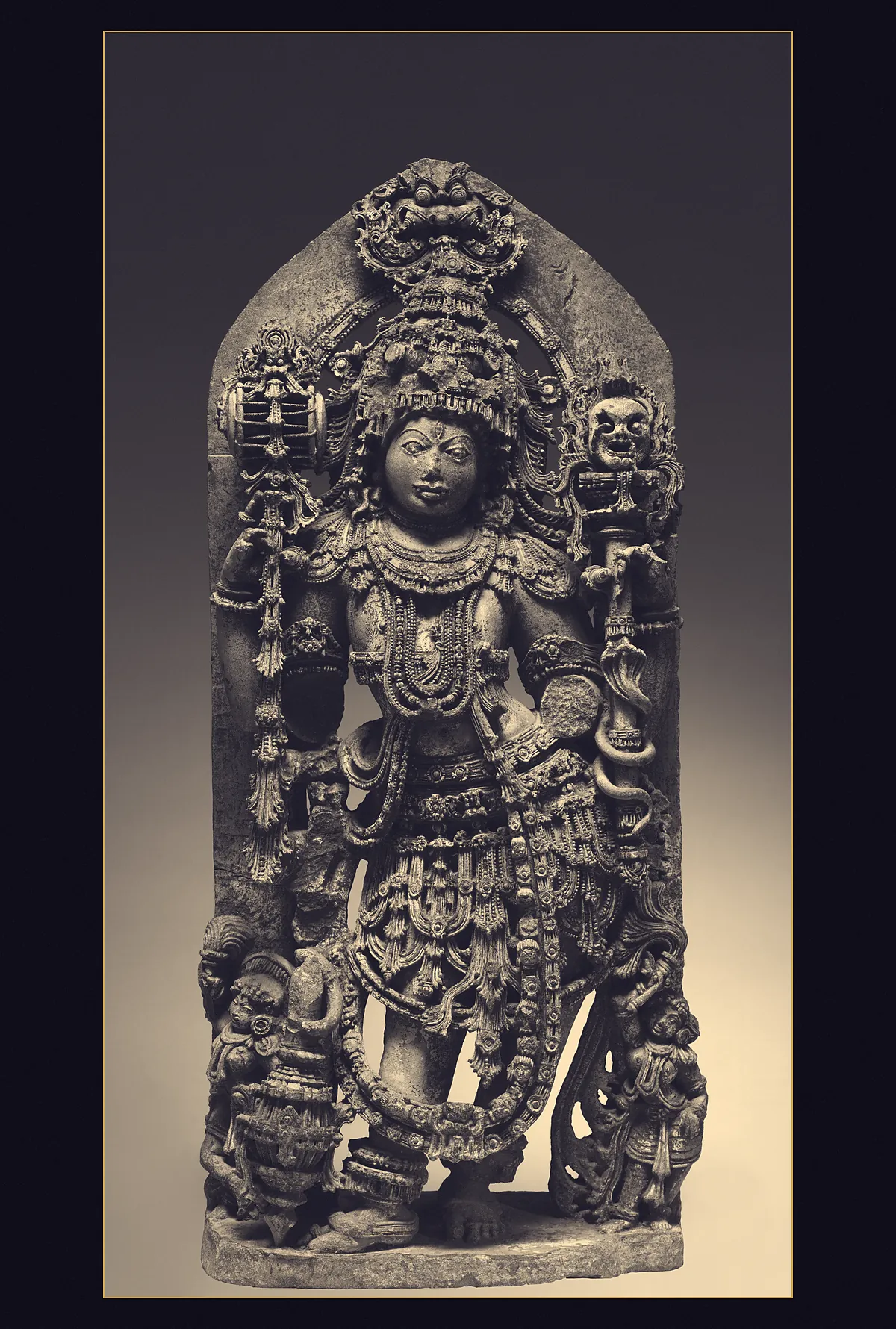 A four-armed Hoysala stone guardian figure standing under a carved arch, holding a drum and a staff, two small attendants at his feet