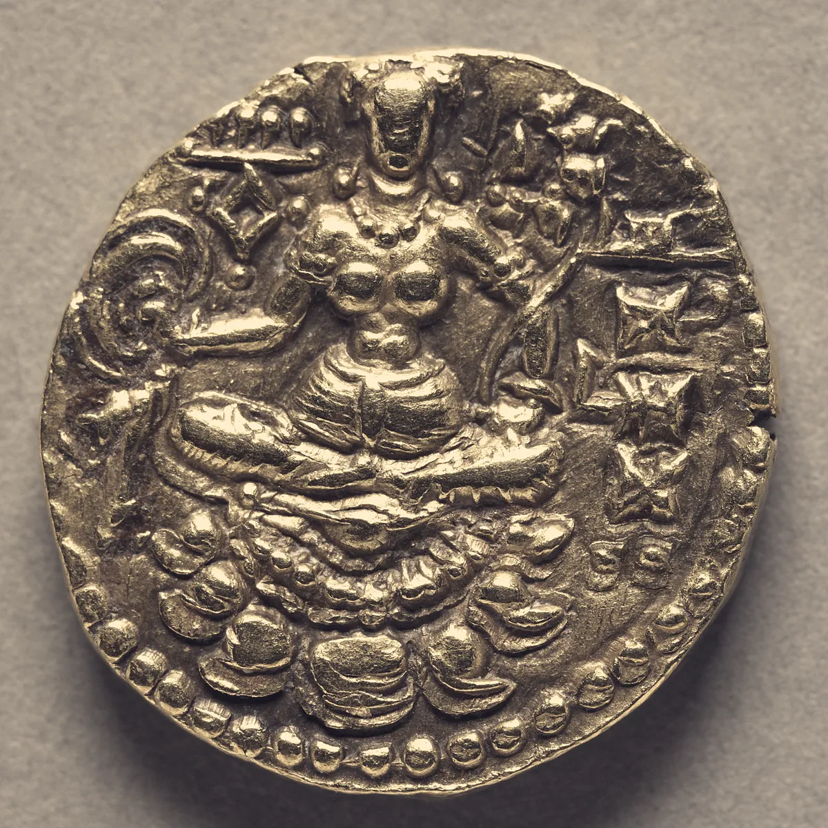 The reverse of a worn Gupta gold coin showing a nimbate goddess seated cross-legged on a lotus, holding a fillet in her raised hand and a long stem in the other, with a Brahmi legend in a column at the right and a dotted border round the flan.