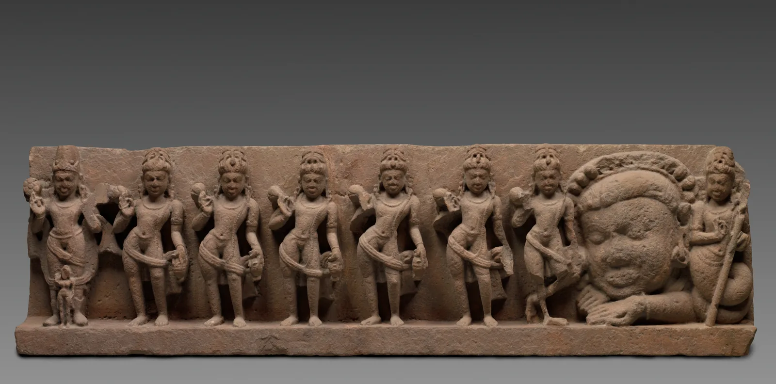 A long sandstone lintel carved with the nine grahas in a row: the Sun with Dawn at his feet, five near-identical standing figures, Saturn on one leg, Rāhu as a great bodiless head, and a serpent-tailed figure at the end